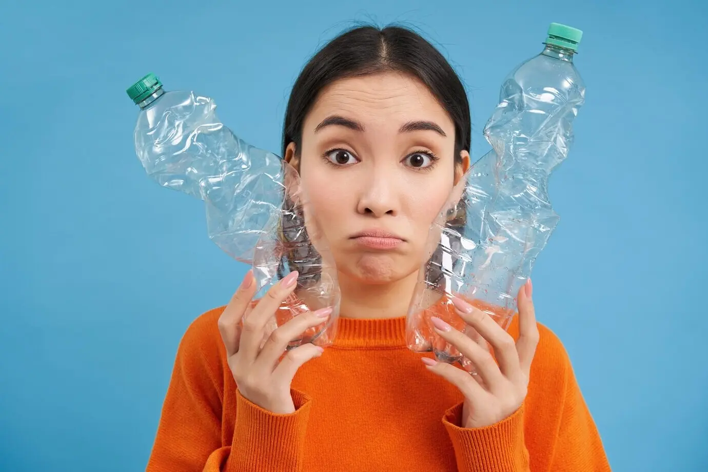Porträt einer traurigen asiatischen Frau mit Plastikflaschen, verärgert über den Mangel an Recyclingzentren, vor blauem Hintergrund.